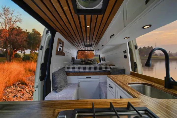 Modern rv rental interior features a slatted wood ceiling, built-in kitchen with sink & stovetop, a comfy bed, and nature views.. Mercedez Sprinter 2015