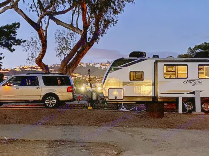 An RV rental: silver SUV hitched to a Grand Design Imagine travel trailer at dusk, lights on, ready for camping.. Grand Design Imagine 2021