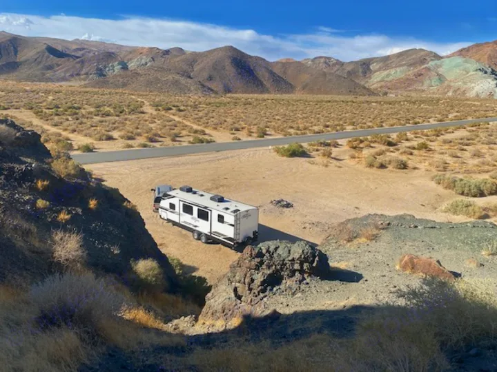An RV rental travel trailer parked in a sandy desert spot, surrounded by arid brush, with mountains and a road in the distance.. Grand Design Imagine 2021