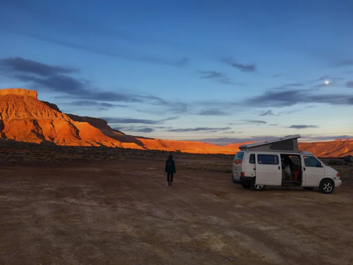 White pop-top RV rental camper van at desert sunset, person walking, moon visible. Ideal outdoor adventure.. Volkswagen Eurovan Full Camper 2001