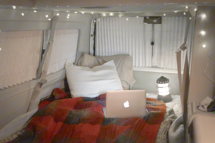 Cozy RV rental interior: bed with plaid blanket, laptop, and lantern, lit by ambient string lights, offering comfort on the go.. Volkswagen Eurovan Full Camper 2001