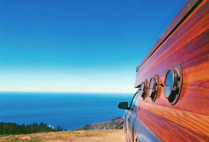 This unique RV rental, a custom wooden van with porthole windows, overlooks the ocean from a cliff under a clear blue sky.. Custom Camper 2017