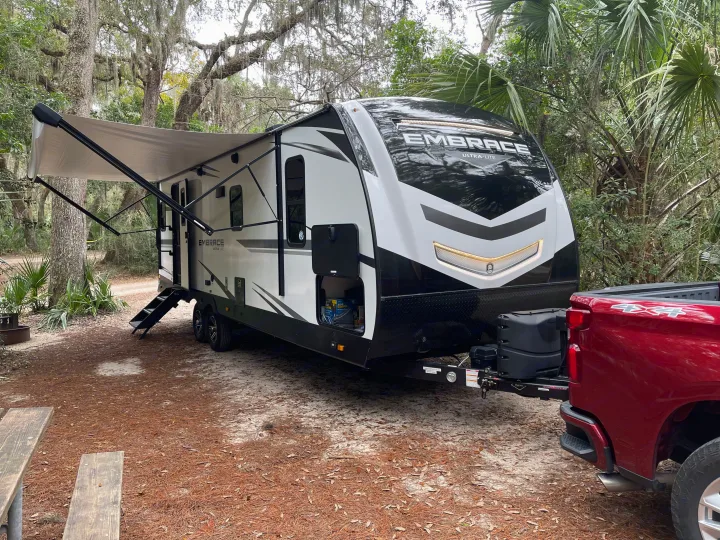RV rental setup: A white travel trailer with an extended awning and red truck parked in a wooded campsite.. Cruiser RV Embrace Ultra Lite 2022