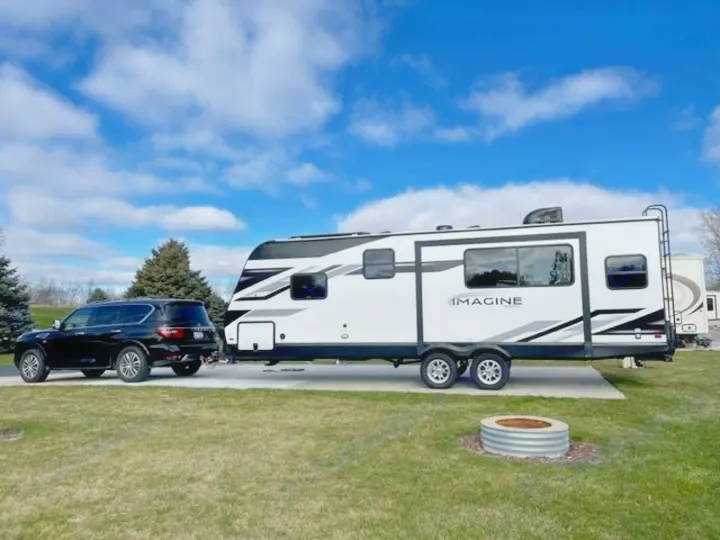 A black SUV is perfectly hitched to a white Imagine travel trailer, set up on an RV rental site with a fire pit.. Grand Design Imagine 2022
