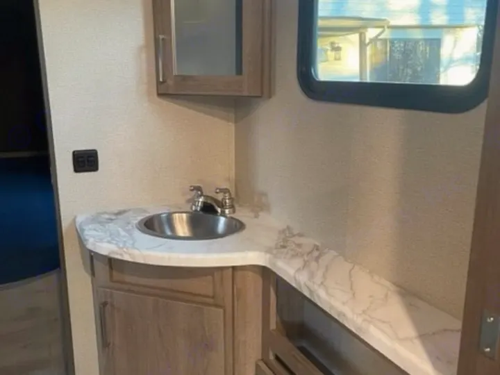 Interior of an RV rental bathroom, showing a compact vanity with sink, faucet, upper cabinet, and window.. Grand Design Imagine 2022