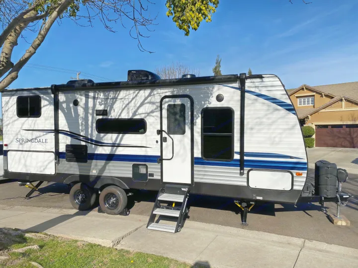 A white and blue Springdale travel trailer, perfect for an RV rental. Parked with steps down under tree shadows.. Keystone Springdale 2021