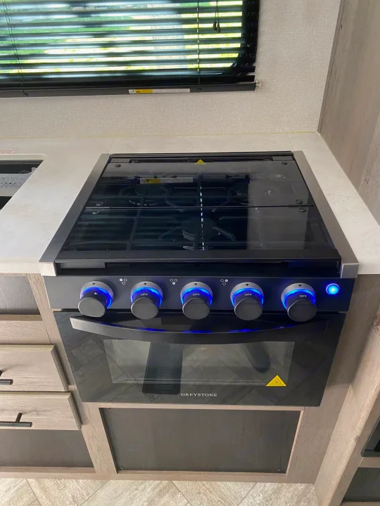 An RV rental kitchen with a sleek Greystone gas range and oven. The black cooktop features blue illuminated control knobs.. Keystone Springdale 2021