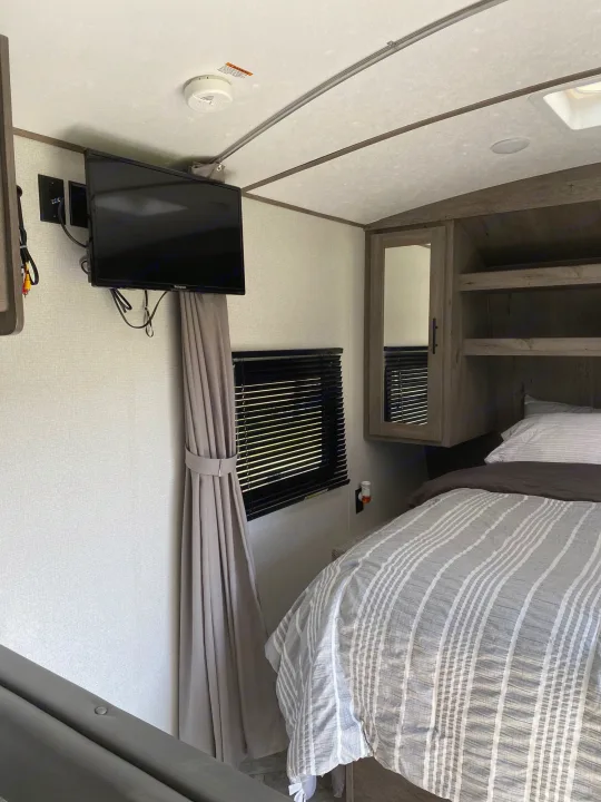Cozy interior of an RV rental bedroom featuring a bed, mounted TV, window with blinds, and cabinet.. Keystone Springdale 2021