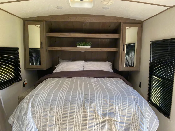 This cozy RV rental bedroom features a comfy bed with striped bedding, built-in wood shelving, mirrored cabinets, and blackout blinds.. Keystone Springdale 2021