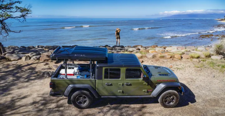 Green Jeep Gladiator, equipped for RV rental adventures with rooftop tent, by the ocean. Person enjoys the scenic view.. Jeep Gladiator Rubicon 2023
