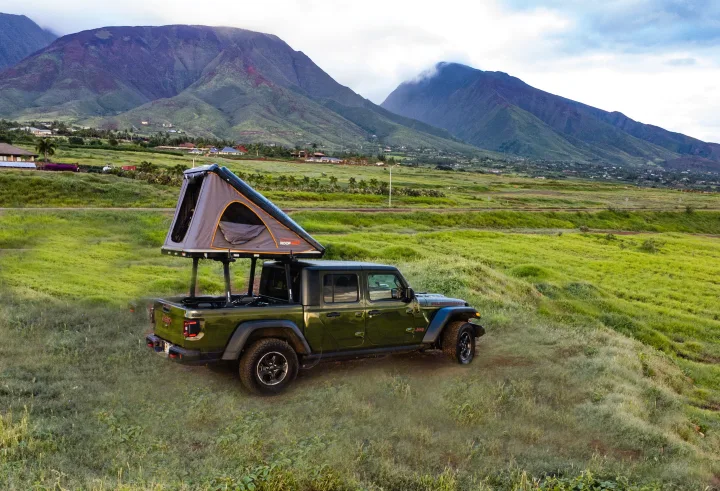 Ready for your RV rental adventure? A green Jeep Gladiator with a mounted rooftop tent sits on a grassy hill, backed by misty mountains.. Jeep Gladiator Rubicon 2023