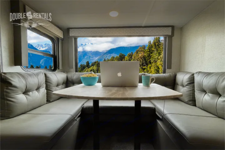 Inside an RV rental: a spacious dinette with gray seating, a laptop, popcorn, and an epic mountain view through large windows.. Forest River Wildwood X-Lite 2020