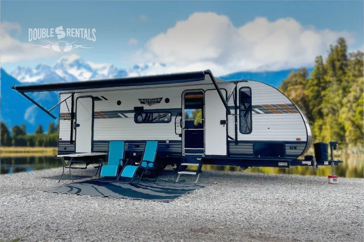 Experience nature with Double S Rentals' travel trailer RV rental, featuring an awning, chairs, and mat by a scenic lake.. Forest River Wildwood X-Lite 2020
