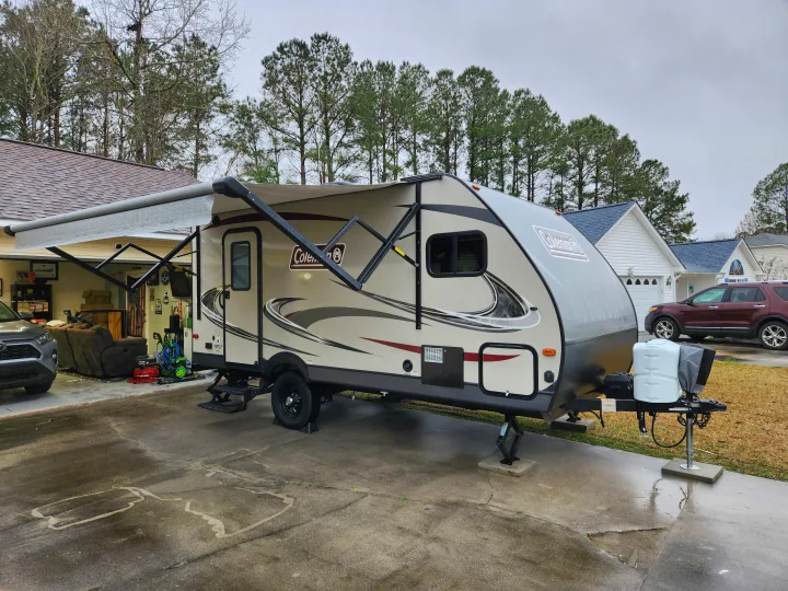 A beige Coleman travel trailer, possibly an rv rental, is parked on a wet driveway with its large awning extended.. Dutchmen Coleman Light LX 2020
