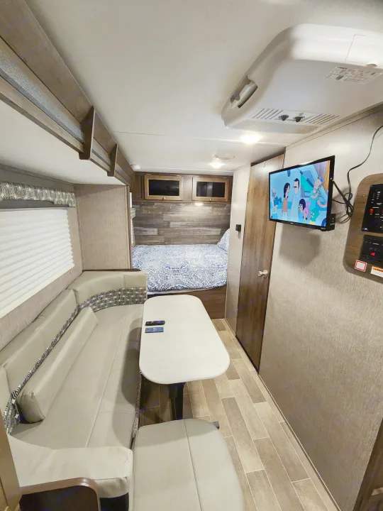 Interior view of an RV rental showing a compact bedroom, dining area, and TV playing a cartoon.. Dutchmen Coleman Light LX 2020