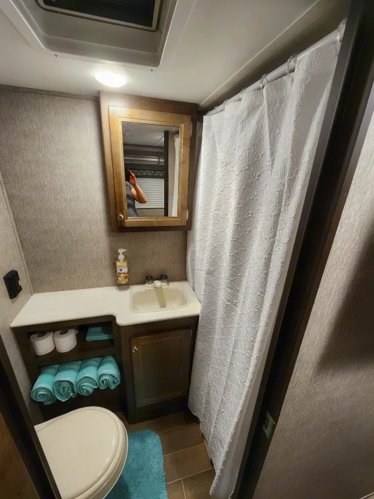 This RV rental bathroom features a compact shower, sink, toilet, mirrored vanity, & organized storage.. Dutchmen Coleman Light LX 2020