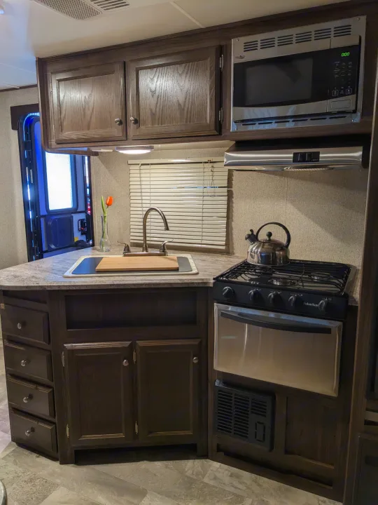 This rv rental kitchen features dark wood cabinets, a compact stove, microwave, and sink, showing a functional and modern interior.. Coachmen Apex LE 2018