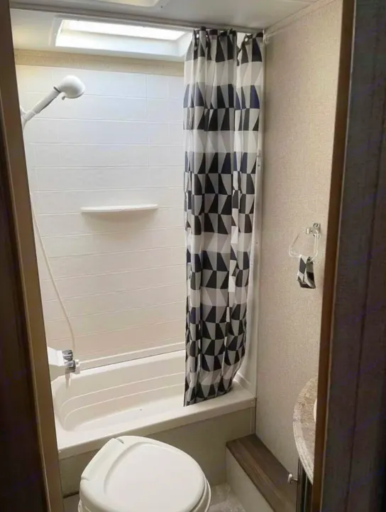 Compact RV rental bathroom features a clean shower/tub combo, toilet, and vanity with a patterned curtain and skylight.. Coachmen Apex LE 2018
