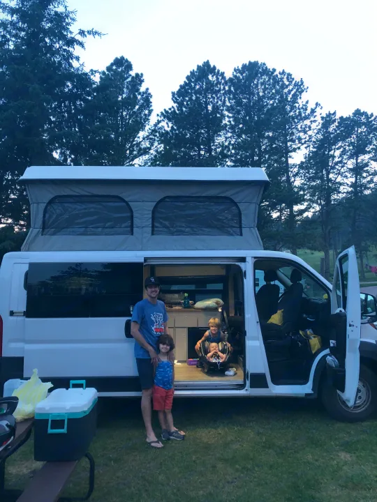 A family of four enjoys their RV rental camper van with a pop-top, parked on grass at a scenic campsite.. Dodge Promaster 2017