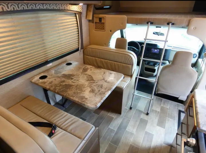 This RV rental interior shows a dinette, window, and an overhead bunk with a ladder leading to the cab.. Thor Magnitude 2021