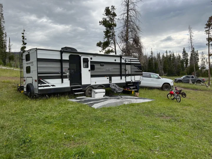 RV rental parked in a grassy field with forest backdrop under cloudy sky, complete with outdoor mat & bikes.. Grand Design Transcend Xplor 2022