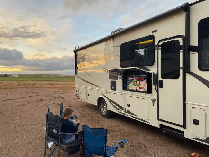 An RV rental provides outdoor entertainment, as a boy watches basketball on its pull-out TV amidst a scenic, open landscape.. Jayco Alante 2021