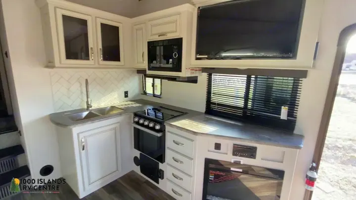An RV rental kitchen, featuring a sink, microwave, oven, TV, and stylish white cabinets with herringbone backsplash.. Keystone Cougar 2022