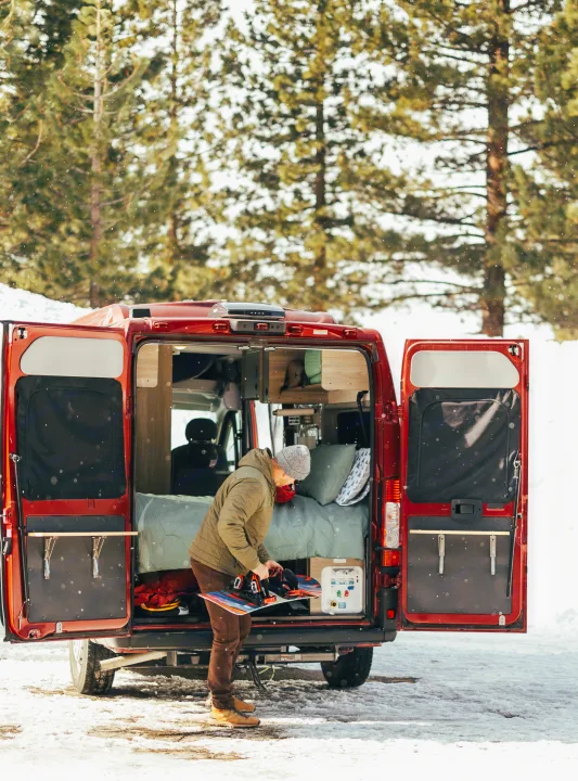 An RV rental enables winter adventures. A person readies their snowboard at the open back of a red campervan amidst snowy trees.. Solis 59P Solis 2022