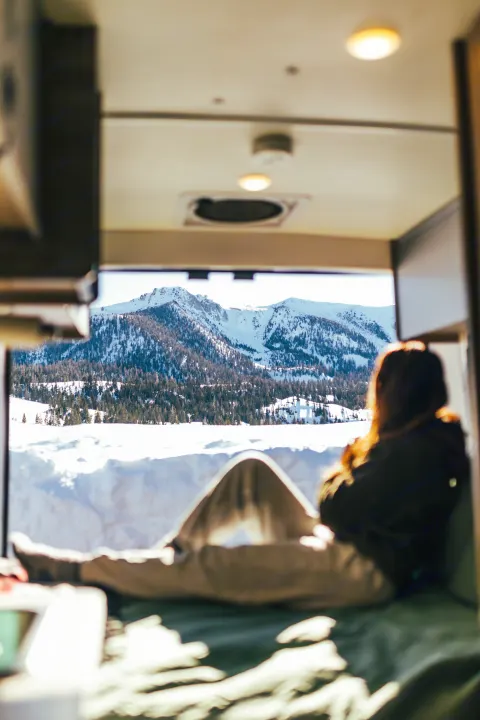 Enjoying a serene winter mountain view from inside an RV rental. A person relaxes on a bed, looking out at the snowy peaks and forests.. Winnebago Solis 2022