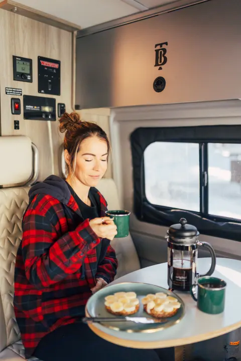 A woman in a cozy plaid shirt sips coffee and eats banana toast inside an RV rental, ready for travel adventures.. Winnebago Solis 2022