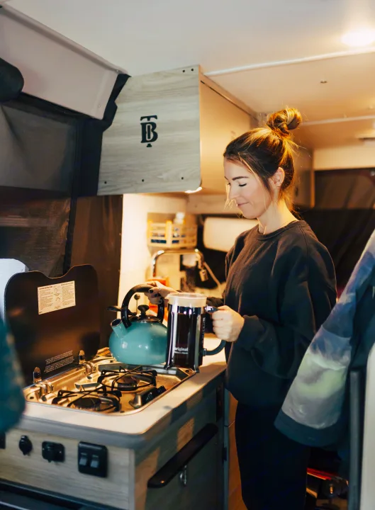 Woman prepares coffee with a French press in a bright rv rental kitchen. Kettle boils on the stove.. Winnebago Solis 2022