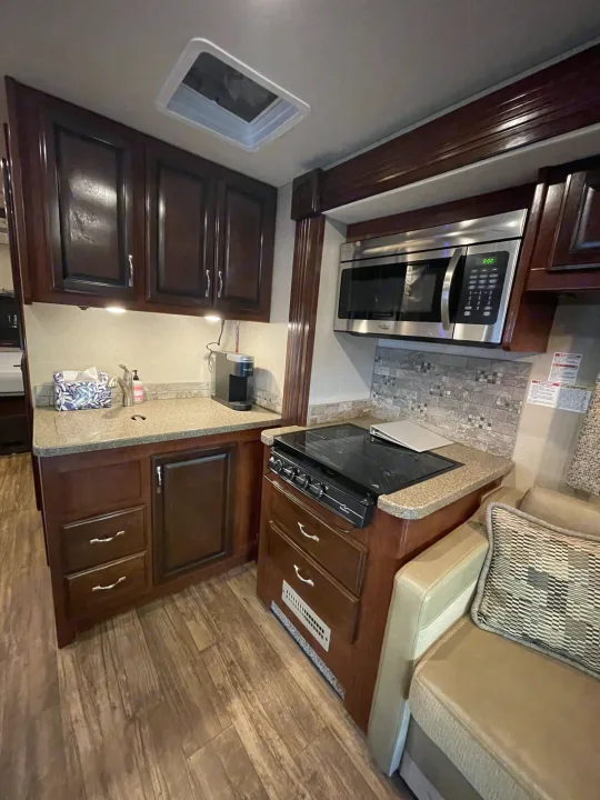 RV rental kitchen with dark wood cabinets, stainless microwave/stovetop, sink, and wood-look flooring, beside a cozy seating area.. Jamboree Jamboree Motorhome 2017