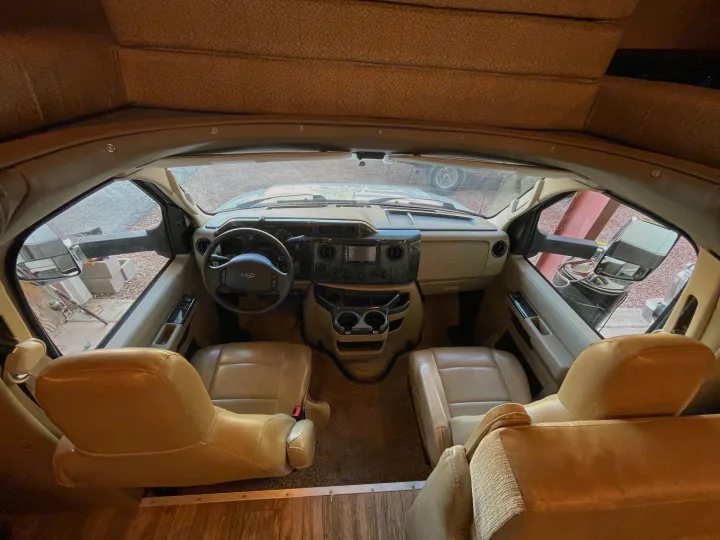 Inside an RV rental, a wide view of the tan-colored cockpit with two front seats, a steering wheel, and dashboard.. Jamboree Jamboree Motorhome 2017