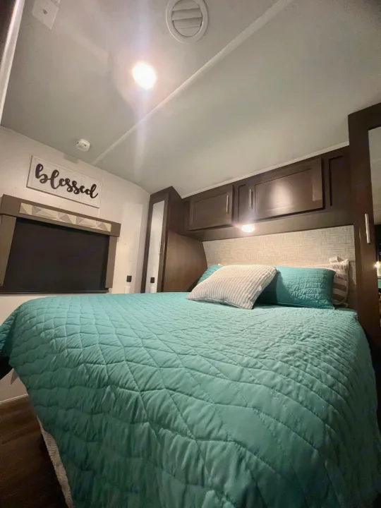 Inviting RV rental bedroom with a teal quilted comforter, comfy pillows, overhead cabinets, and a "Blessed" wall sign.. Forest River Wildwood 2020