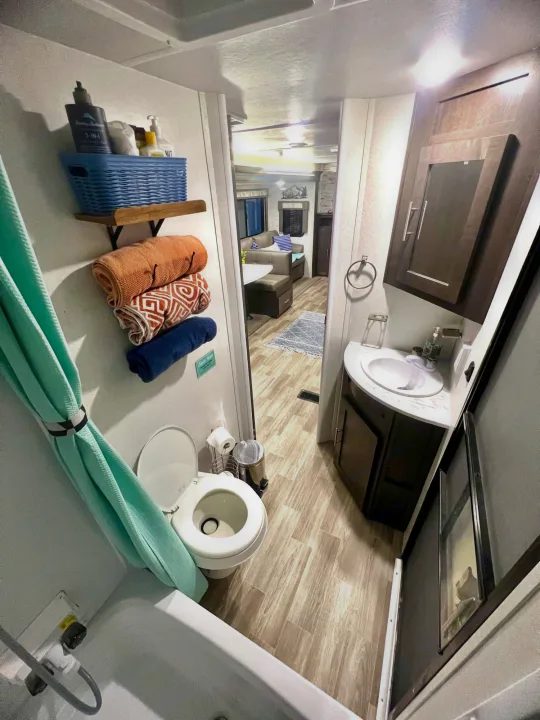 Spacious RV rental bathroom: toilet, shower with teal curtain, vanity. Storage shelf with towels. View into living area.. Forest River Wildwood 2020