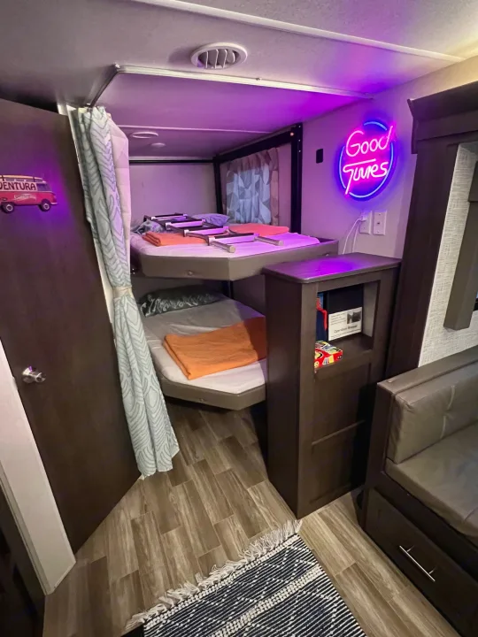 This RV rental offers a compact and cozy sleeping area with bunk beds, an adjacent sofa, and a colorful neon sign.. Forest River Wildwood 2020
