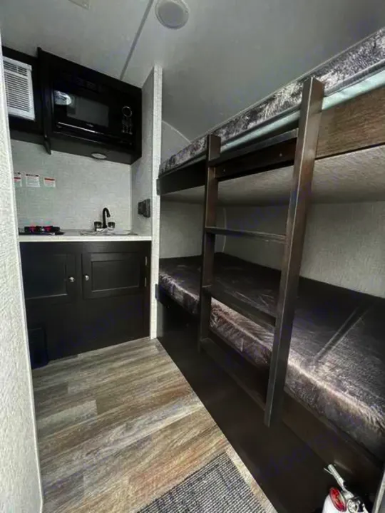 Interior of an RV rental showing a compact kitchen area with microwave/sink and two bunk beds with a ladder for sleeping.. Braxton Creek Bushwhacker Plus 2021