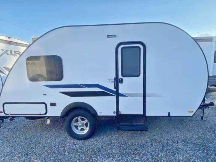 Side view of a white, compact teardrop RV rental. This travel trailer, marked 17 BH, has dark blue graphics & off-road tires.. Braxton Creek Bushwhacker Plus 2021