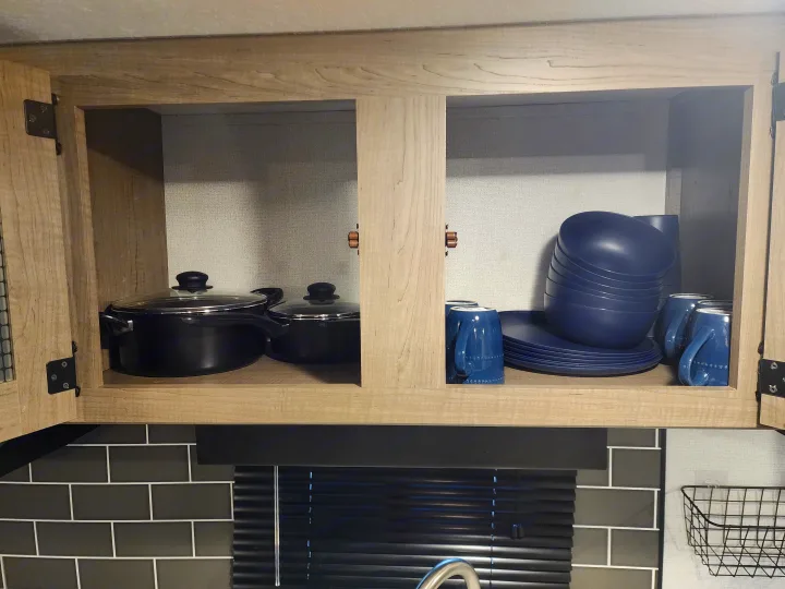Open light wood RV rental kitchen cabinets reveal organized black pots, blue plates, bowls, and mugs on shelves.. Keystone RV Hideout 2022