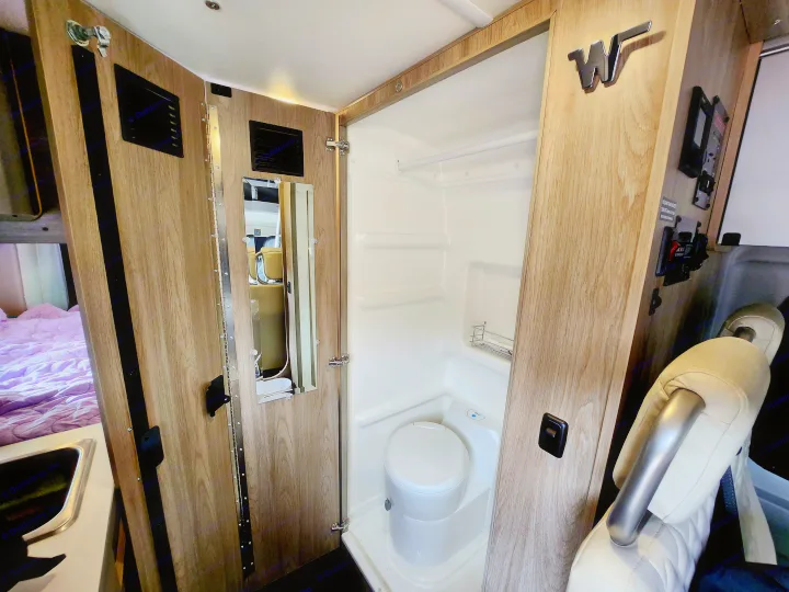 Interior of an rv rental, showing a compact bathroom with toilet and shower stall. A bed and small sink are to the left.. Winnebago Solis 2023