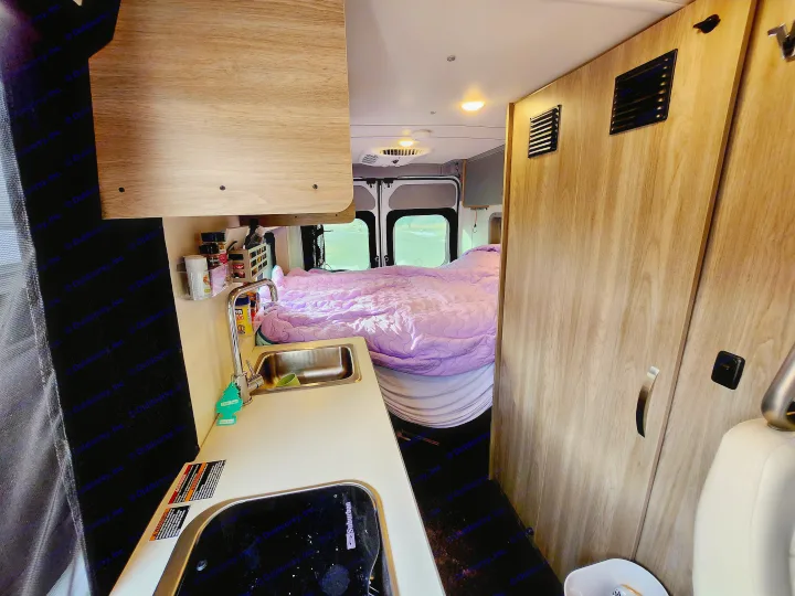 Cozy rv rental interior showing a compact kitchen with sink, stove, and a comfortable bed in the back of the camper van.. Winnebago Solis 2023