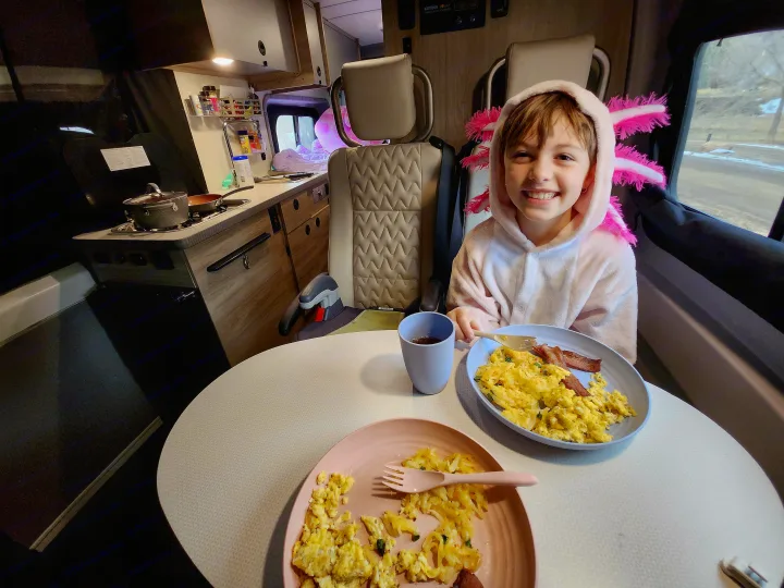A happy child in a pink axolotl robe eats eggs and bacon for breakfast at a cozy table inside an RV rental.. Winnebago Solis 2023