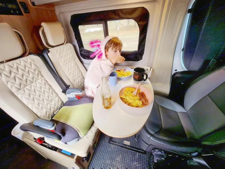 Child in a pink axolotl robe eats breakfast at a table in an RV rental, a cozy scene for family travels.. Winnebago Solis 2023