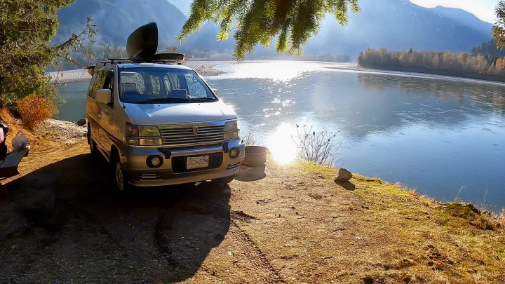 RV rental adventure! A silver camper van with a kayak on its roof is parked by a sparkling river with mountains.. nissan elgrand 1997