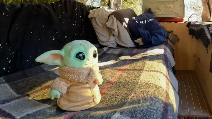 Grogu plush on a blanket in a sunlit RV rental interior, featuring bedding and wood panels for a cozy travel setup.. nissan elgrand 1997