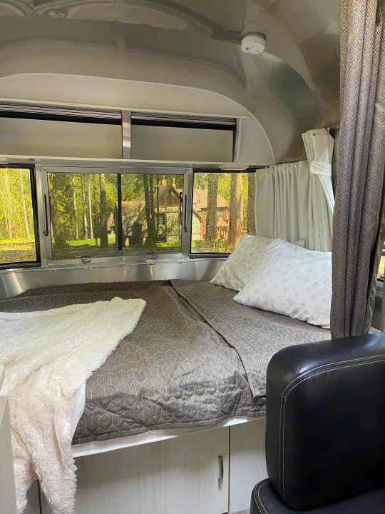 RV rental bedroom interior: cozy bed with grey quilt, white blanket, pillows, and window views of lush trees and a home.. Airstream Sport 2019