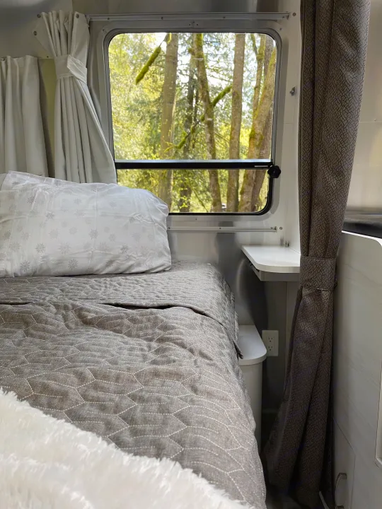 Cozy RV rental bed with grey quilted comforter and white pillow, alongside a window view of lush trees and curtains.. Airstream Sport 2019