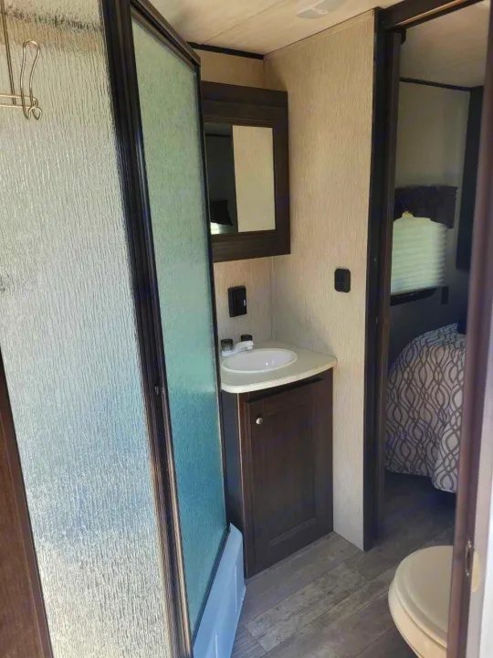 This RV rental features a compact bathroom with a textured glass shower, sink vanity, and partial view of the bedroom.. Heartland RVs Pioneer 2020