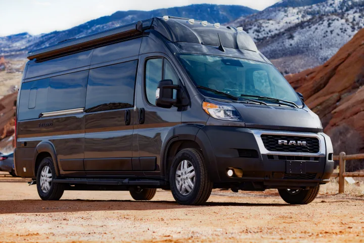 Dark gray RV rental camper van on a dirt path. Snow-capped mountains and red rock formations create a scenic backdrop.. Thor Tellaro 2023