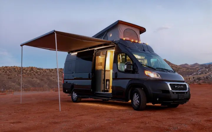 Dark grey camper van RV rental at dusk, with pop-top & awning extended on red dirt. Illuminated interior invites adventure.. Thor Tellaro 2023